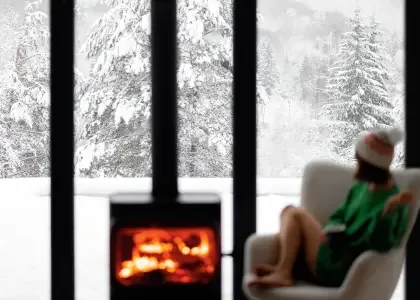 photo d'une femme sur un fauteuil à côté d'un foyer de feu face à une grande fenêtre avec la vue sur une forêt enneigée